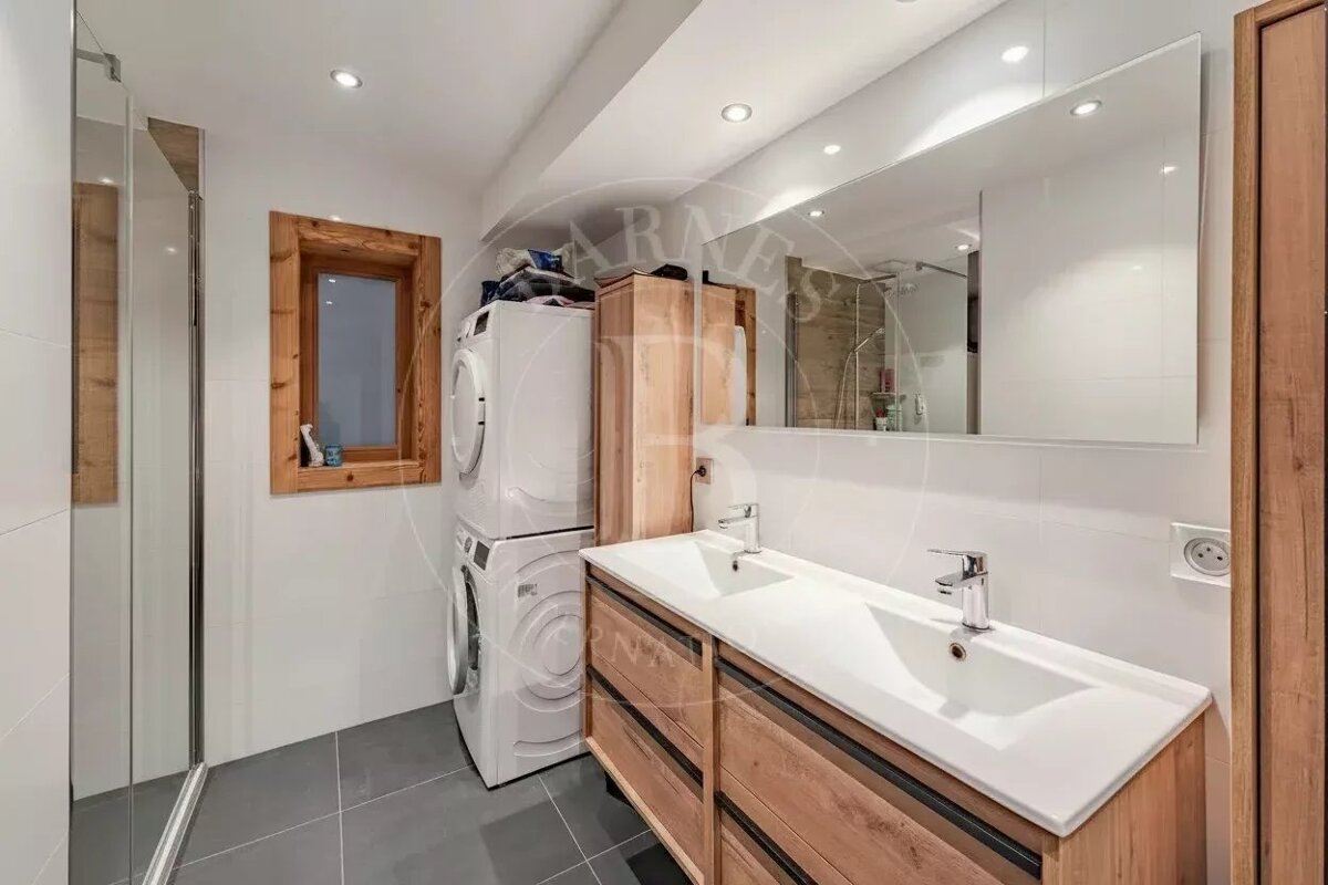 Modern bathroom featuring a wooden double vanity, stacked washer and dryer, a large mirror, and a glass shower cabin.