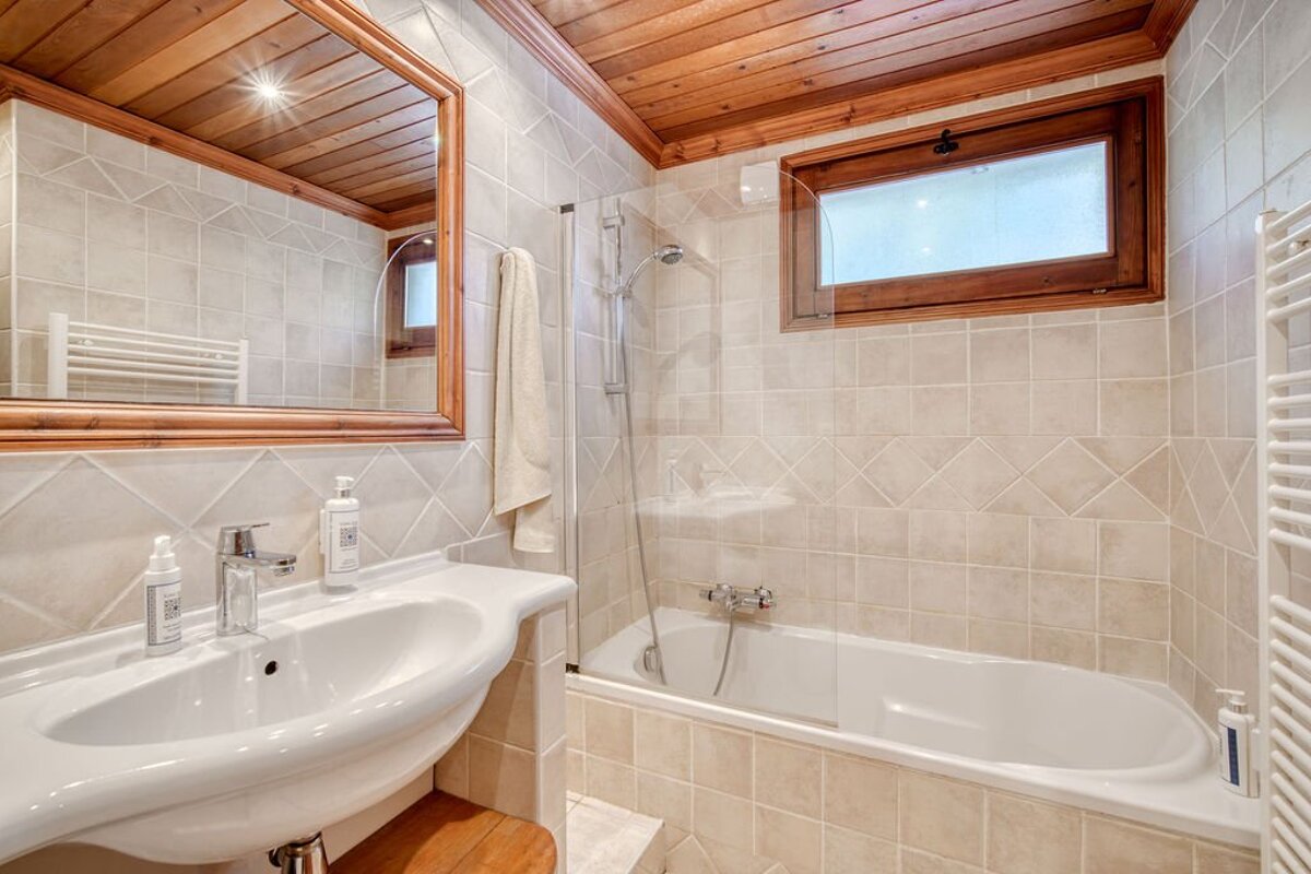 Tiled bathroom with a wooden ceiling, featuring a white sink, large mirror, and a bathtub with a glass shower partition.