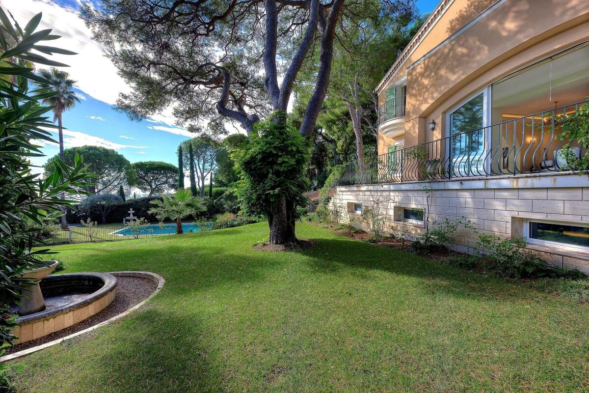 Luxurious villa with a sprawling green lawn, large trees, a swimming pool, and a fountain under a blue, cloudy sky.