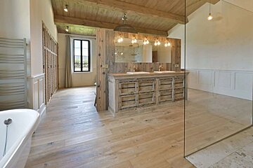 A spacious rustic bathroom with exposed wood beams, light wood floors, a double vanity, a freestanding tub, and a glass shower.