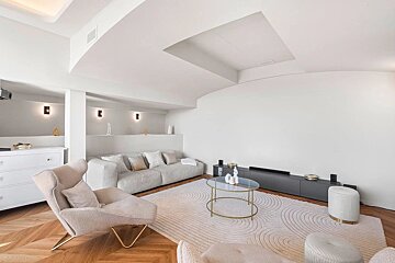 A bright, modern living room with a gray sofa, light armchairs, a unique geometric rug, wood floors, and white curved walls.