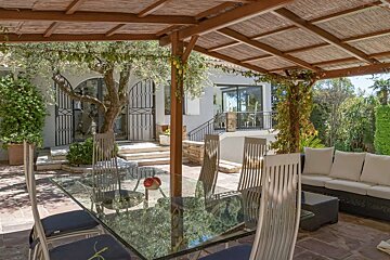 A sun-dappled Mediterranean patio features a glass dining table, outdoor lounge, olive tree, and a modern house under a bamboo pergola.