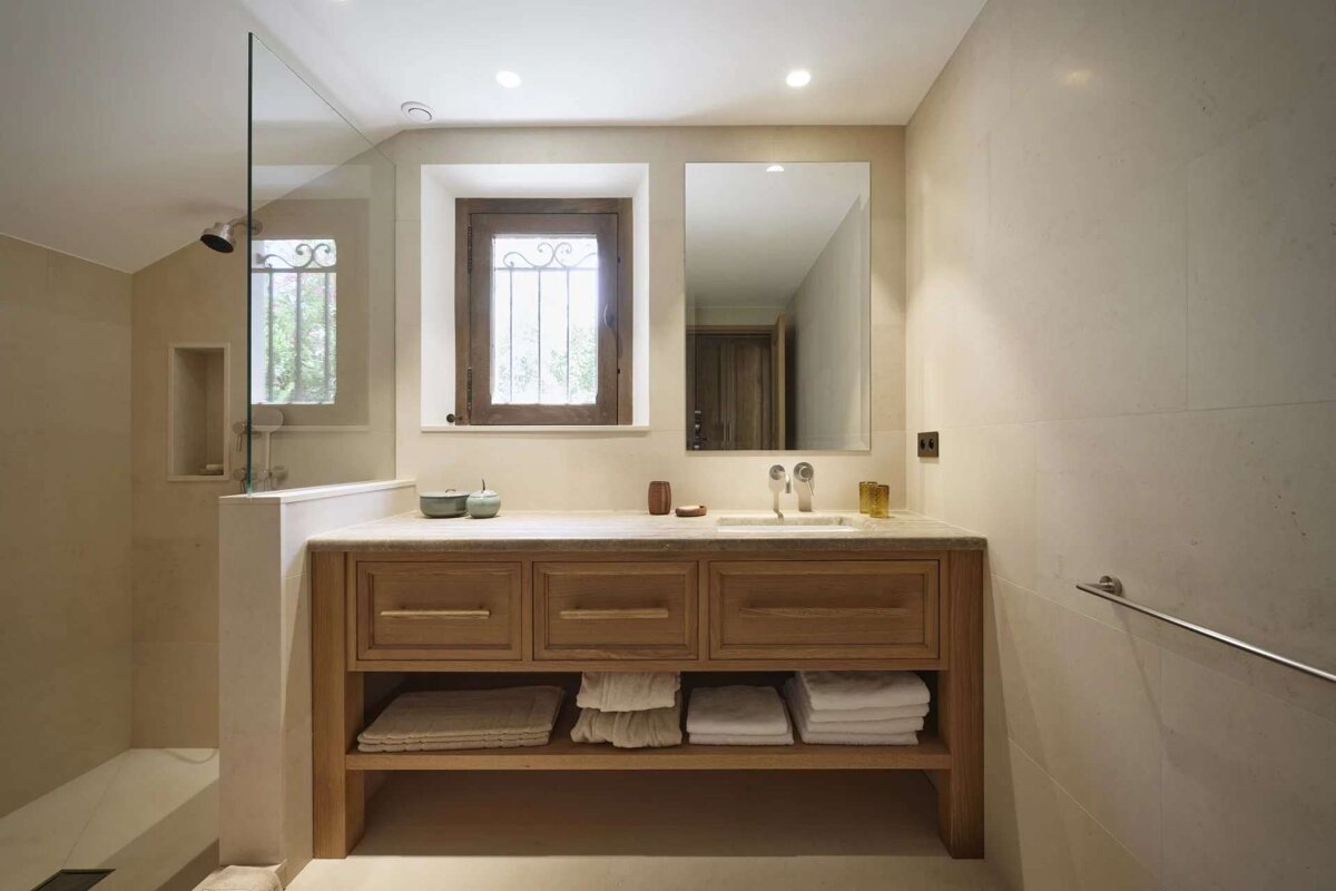 A neutral-toned bathroom features a wooden vanity with a marble top and integrated sink, a large mirror, and a glass-enclosed shower. A window with a decorative iron grille adds character.