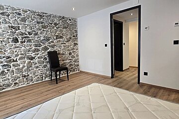 Empty room with a stone accent wall, white walls, wooden floor, black chair, mattress in foreground, and dark doorway leading further in.