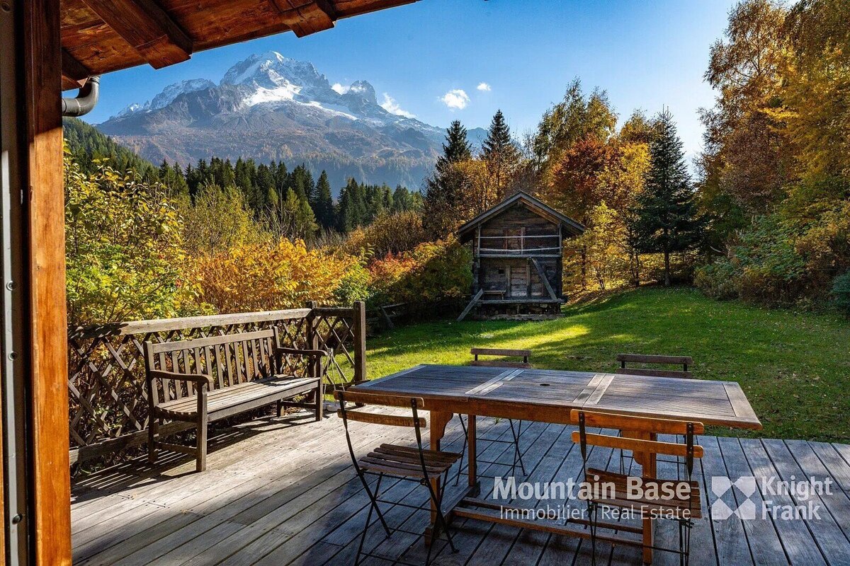 A wooden deck with outdoor furniture overlooks a vibrant autumn mountain landscape. Colorful trees surround a small rustic cabin under a blue sky.
