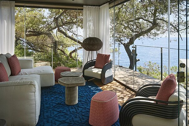 Modern sunroom overlooking the ocean, featuring stylish seating, a blue rug, glass walls, and lush green trees.