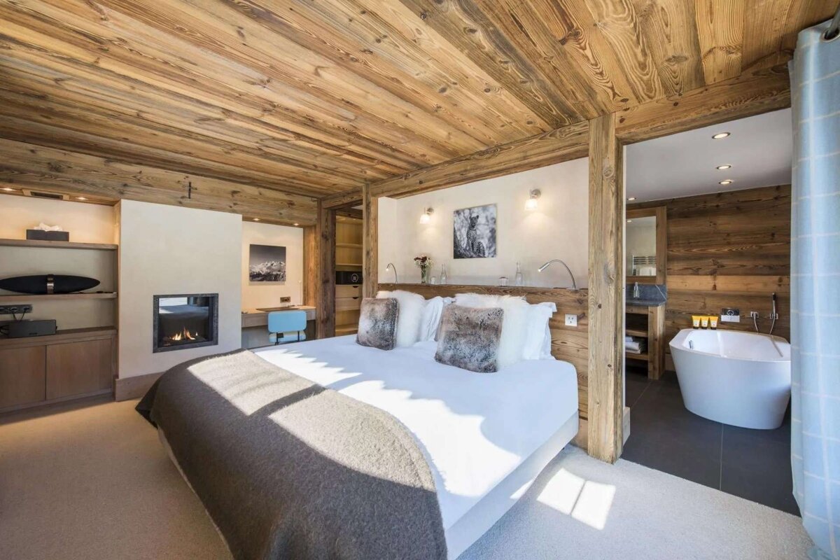 A luxurious, wood-paneled chalet bedroom features a fireplace, a large bed, and an open bathroom with a modern freestanding tub.
