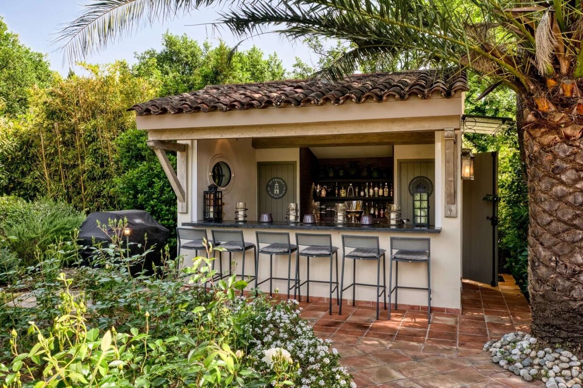An inviting outdoor bar with a tiled roof, bar stools, and a grill, surrounded by lush greenery, a palm tree, and a tiled patio.
