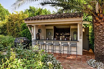 An inviting outdoor bar with a tiled roof, bar stools, and a grill, surrounded by lush greenery, a palm tree, and a tiled patio.