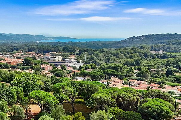 A panoramic view of a coastal town with red-roofed houses nestled in lush green trees, leading to a blue sea and distant hills under a sunny sky.