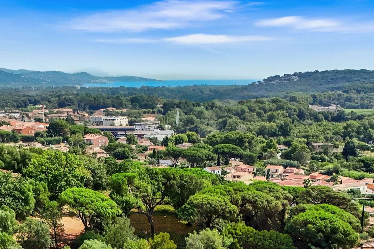 An aerial view of a vibrant green landscape with numerous trees, a town with red-roofed buildings, and a distant blue sea under a clear sky.