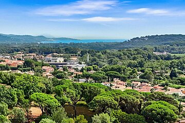 An aerial view of a vibrant green landscape with numerous trees, a town with red-roofed buildings, and a distant blue sea under a clear sky.