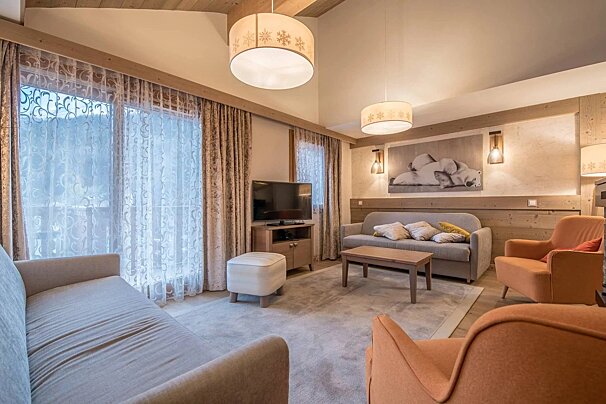 A cozy, rustic-chic living room with grey sofas, orange armchairs, a TV, and large windows, featuring snowflake lights and wooden accents.