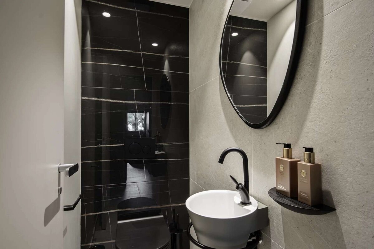 A modern, minimalist washroom with black marble-like tiles, a grey wall, a white sink, black faucet, oval mirror, and a toilet.