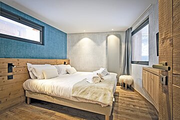 A stylish, modern bedroom with a large bed, wooden headboard, blue accent wall, gray textured walls, and a window. Warm lighting adds coziness.