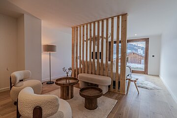 A cozy chalet living space with fuzzy armchairs, wooden tables, and a slat partition dividing it from a bed area. A large window overlooks snowy mountains.