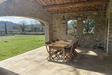 A rustic stone patio with a wooden dining set overlooks a sunny green field and distant trees. A child's blue bike rests by the wall.