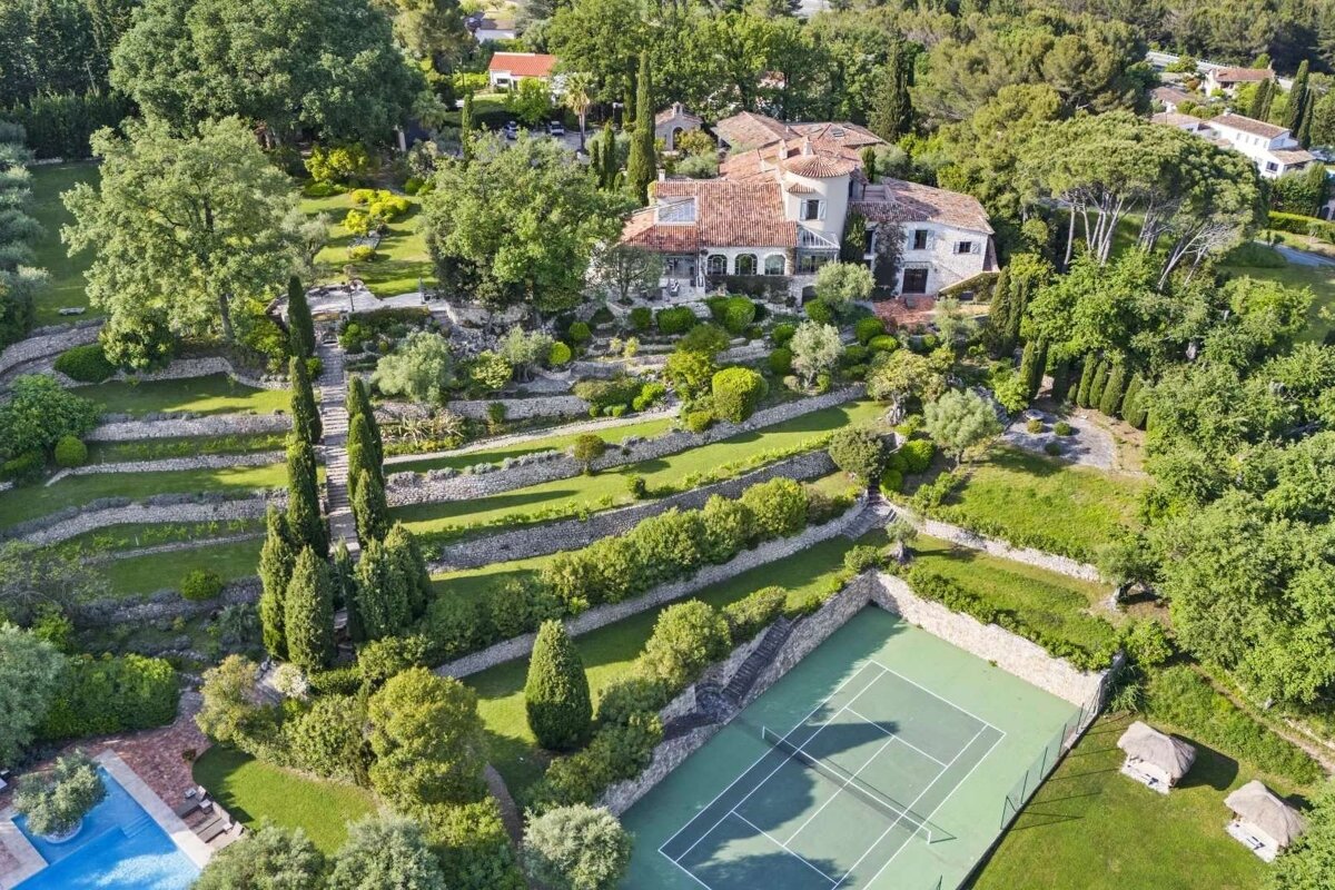 Aerial view of a sprawling Mediterranean estate, featuring a grand house with red roofs, terraced gardens, a tennis court, and a swimming pool.