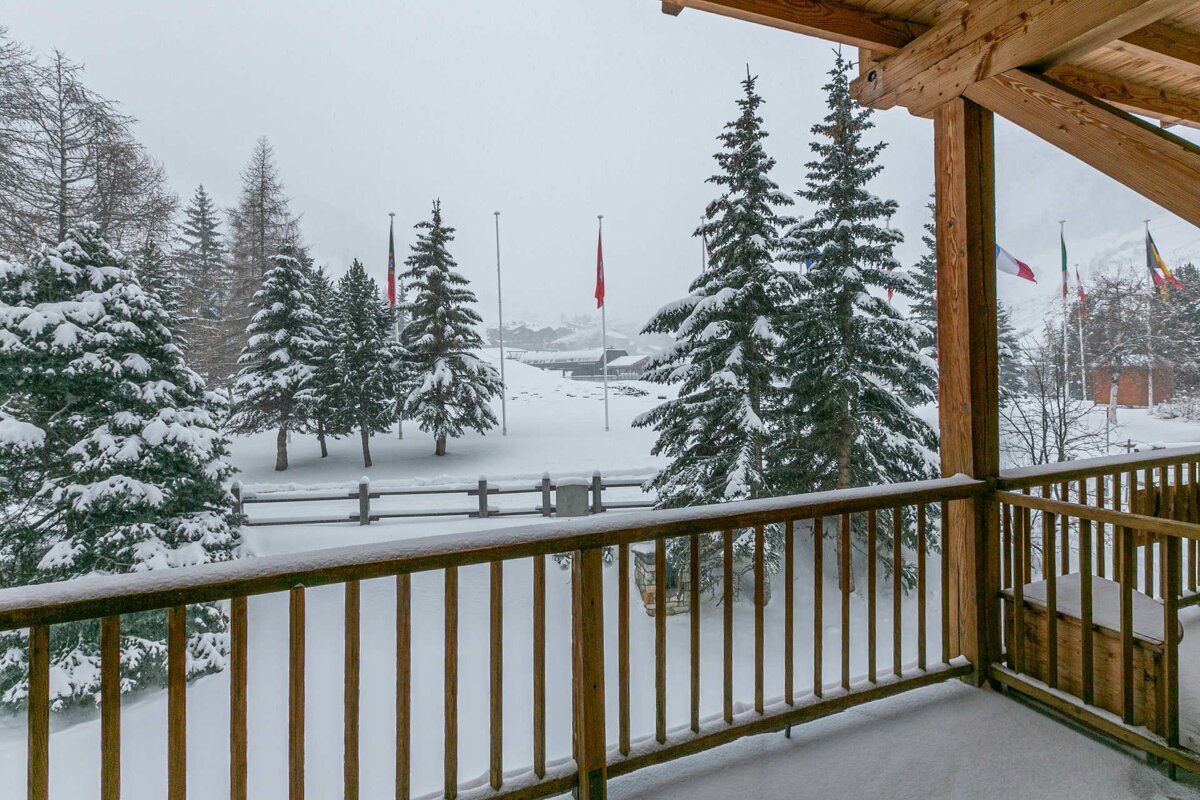 View from a snowy wooden balcony overlooking a snow-covered landscape with pine trees and flags under a white sky.