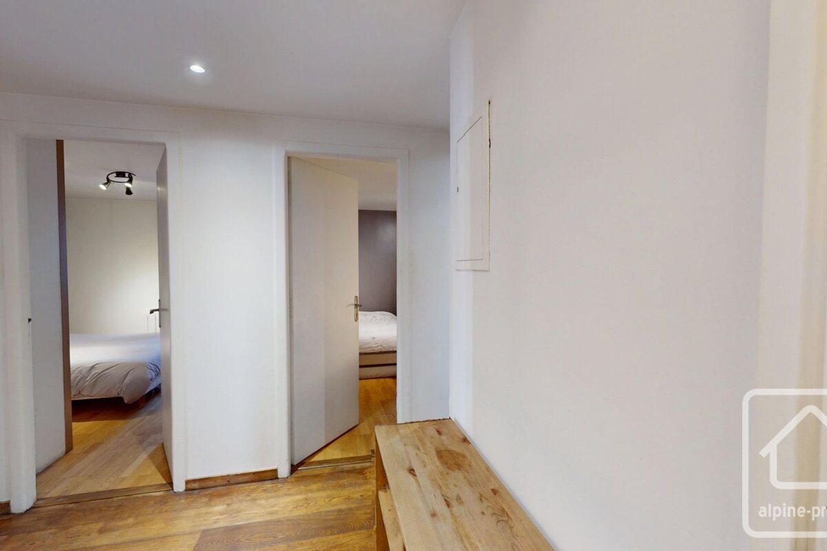 A bright hallway with wooden floors and white walls, featuring two open doors revealing bedrooms and a wooden bench on the right.