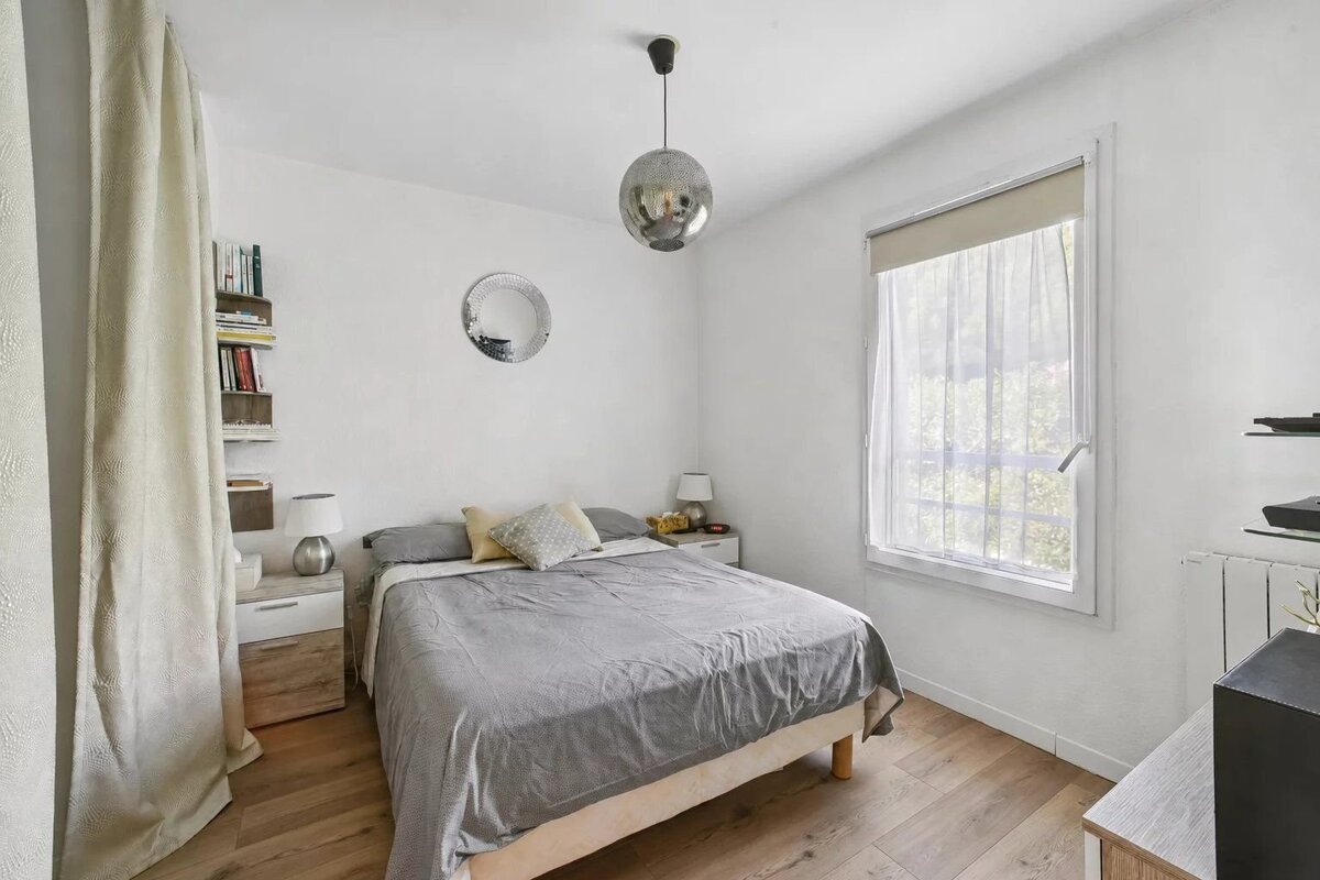 Bright, modern bedroom featuring a bed with grey linens, light wood floors, a large window, and a silver globe pendant light.