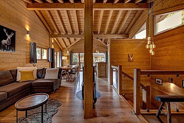 A spacious, warm all-wood chalet interior featuring a living room, dining area, and staircase with modern lighting, creating a cozy, rustic ambiance.