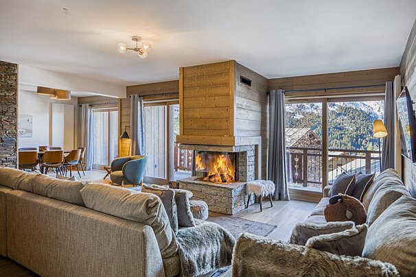 A warm, cozy living room with a lit fireplace, plush sofas, and a dining area, offering a snowy mountain view through large windows.