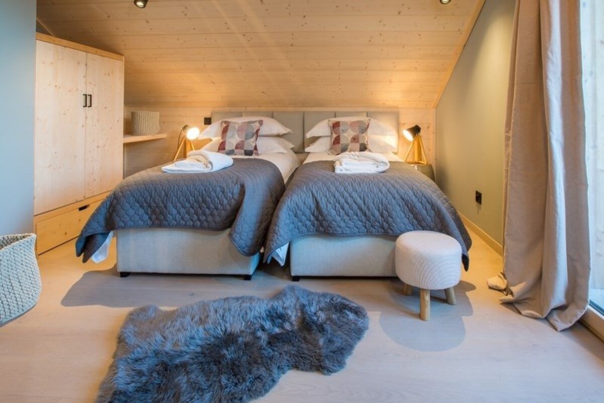 Cozy attic bedroom featuring two twin beds with grey quilts, a slanted wooden ceiling, light wood furniture, and a plush grey rug.