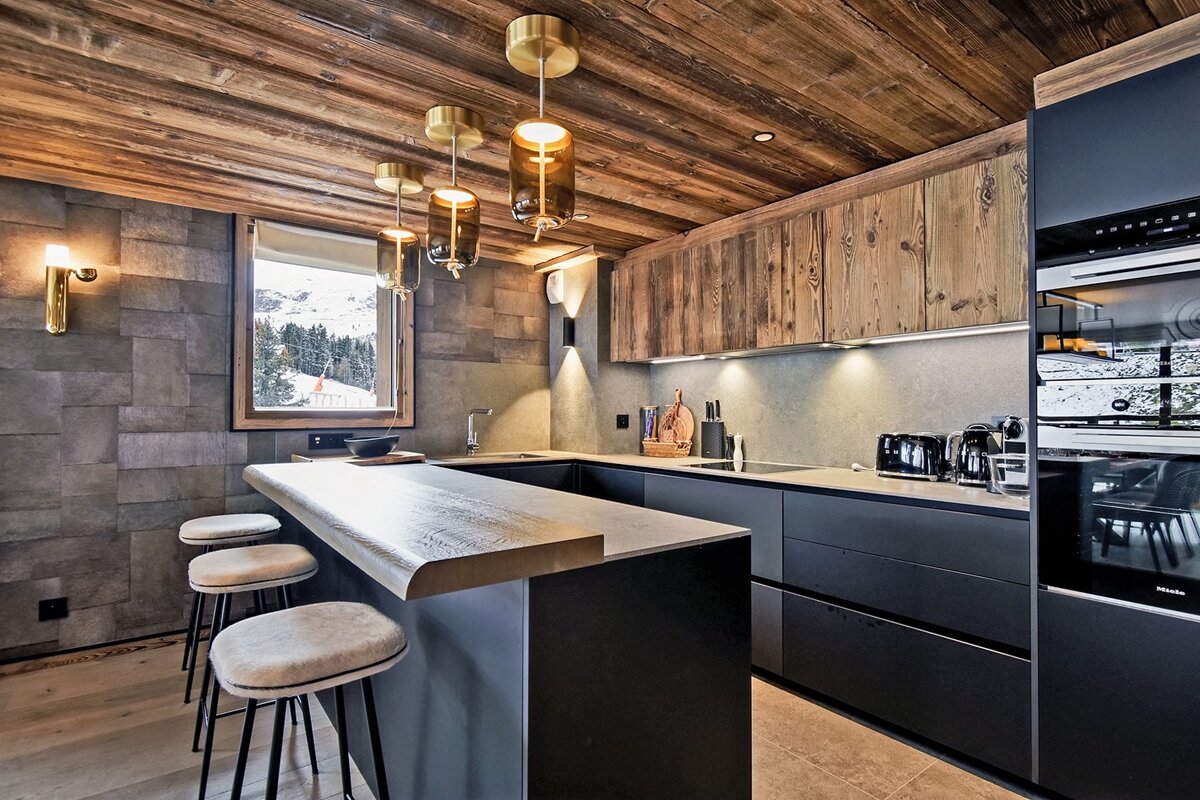 Chic mountain kitchen with dark modern units, rustic wood ceiling & cabinets, bar seating, and a window framing a snowy alpine landscape.
