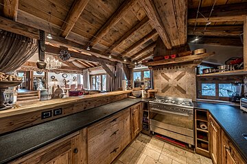 A rustic, open-plan kitchen and living area with rich wooden beams, cabinets, stone backsplash, and a large professional range.