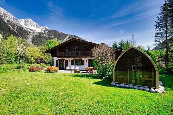 A wooden alpine chalet set on a green lawn with a unique arched pod, backed by snow-capped mountains under a clear blue sky.