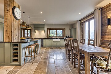 A rustic open-plan kitchen and dining area features sage green cabinets, a large wooden table, and a stone floor. Windows overlook a mountain landscape.
