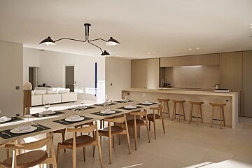 A bright, modern open-plan dining room and kitchen featuring a long wooden table set for guests, a minimalist island bar with stools, and white walls.