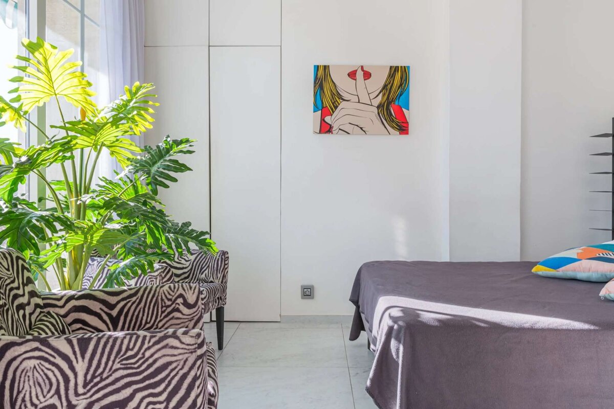 Bright, modern room with a large green plant, zebra print chairs, a pop art shush painting, and a gray bed. Sunlight streams in.