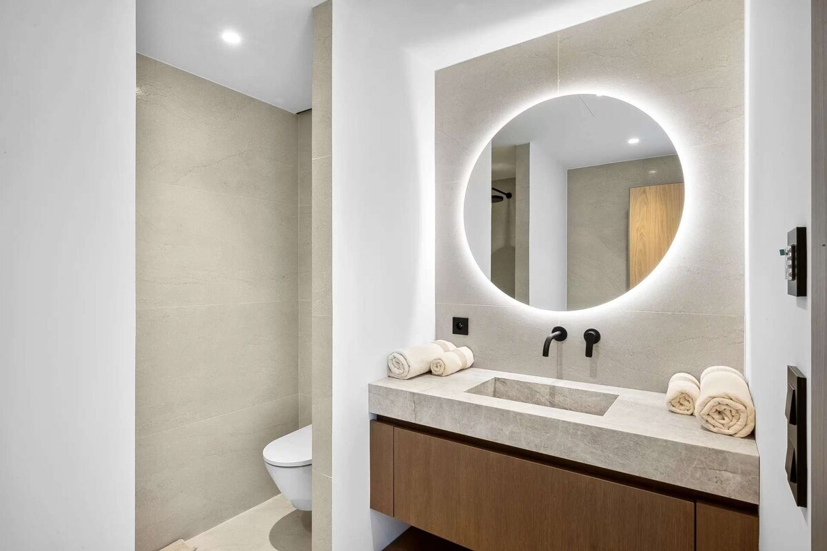 Modern bathroom with illuminated round mirror above a stone vanity, black faucets, and wooden cabinet. Toilet in a tiled alcove.