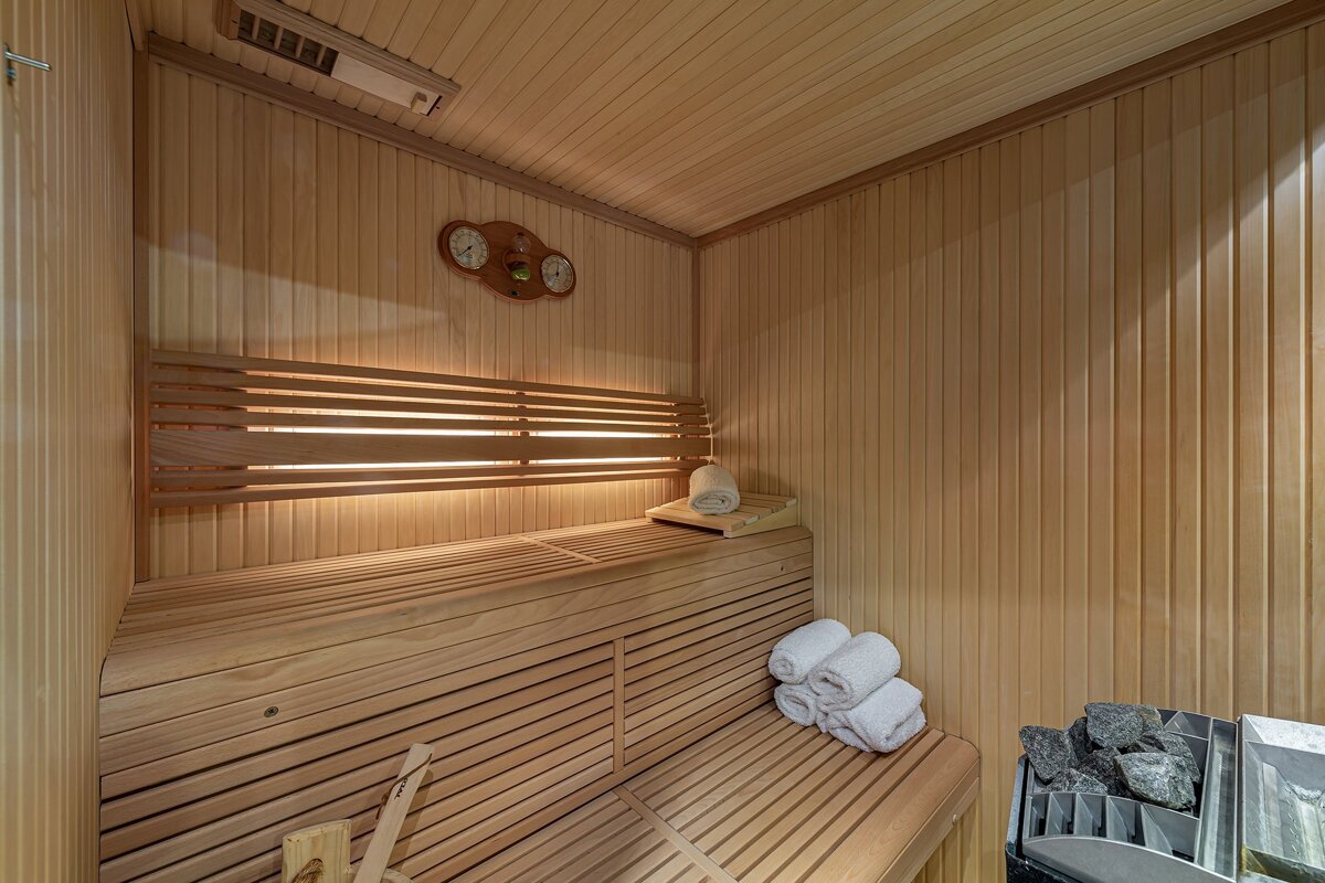 A light wooden sauna interior with multi-level benches, a heater with rocks, white towels, and wall gauges.