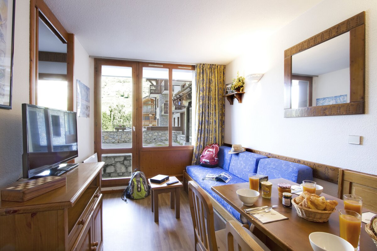 A cozy apartment living/dining area featuring a blue sofa, TV, and breakfast on the table. A large window offers a view of a village outside.