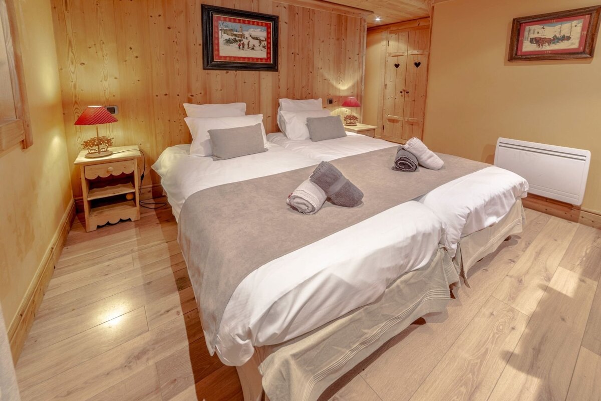 A cozy, rustic bedroom features two single beds, light wood-paneled walls, and a matching floor. Warm lamps illuminate the neutral bedding with folded towels.