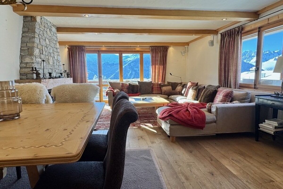 Cozy alpine chalet featuring a wooden dining table, large sectional sofa, stone fireplace, and windows framing snowy mountain views.