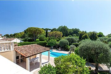 An elegant villa backyard featuring a bright blue swimming pool, a tiled-roof pavilion, and lush green trees under a clear sky.