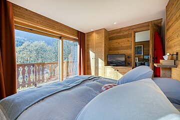 A cozy, wood-paneled bedroom features a large bed, TV, and a stunning balcony view of mountains and a forest under a bright sky.