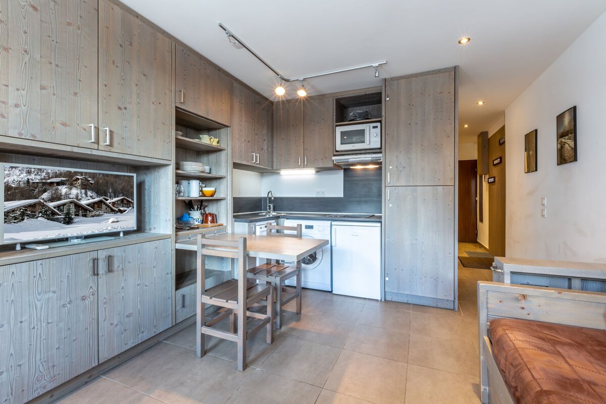 A compact, modern studio with integrated light wood cabinetry, featuring a full kitchenette, dining table, TV area, and sleeping space.