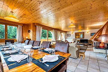 Cozy alpine chalet interior with a long, set wooden dining table, a living area with a warm fireplace, and pine wood-paneled ceilings.