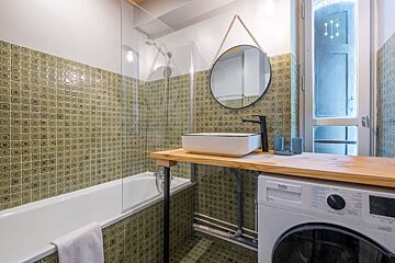 Modern bathroom with green tiles, featuring a bathtub, a wooden counter with a vessel sink, a round mirror, and a washing machine.