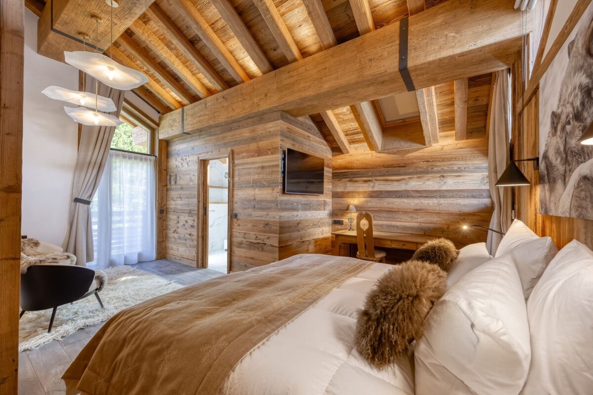 Rustic yet luxurious bedroom with wooden walls, beamed ceiling, a cozy bed, modern lights, and a large window.