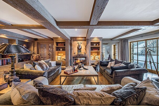 A cozy, luxurious chalet living room with exposed wood beams, a fireplace, plush sofas, and a large window revealing a snowy mountain vista.