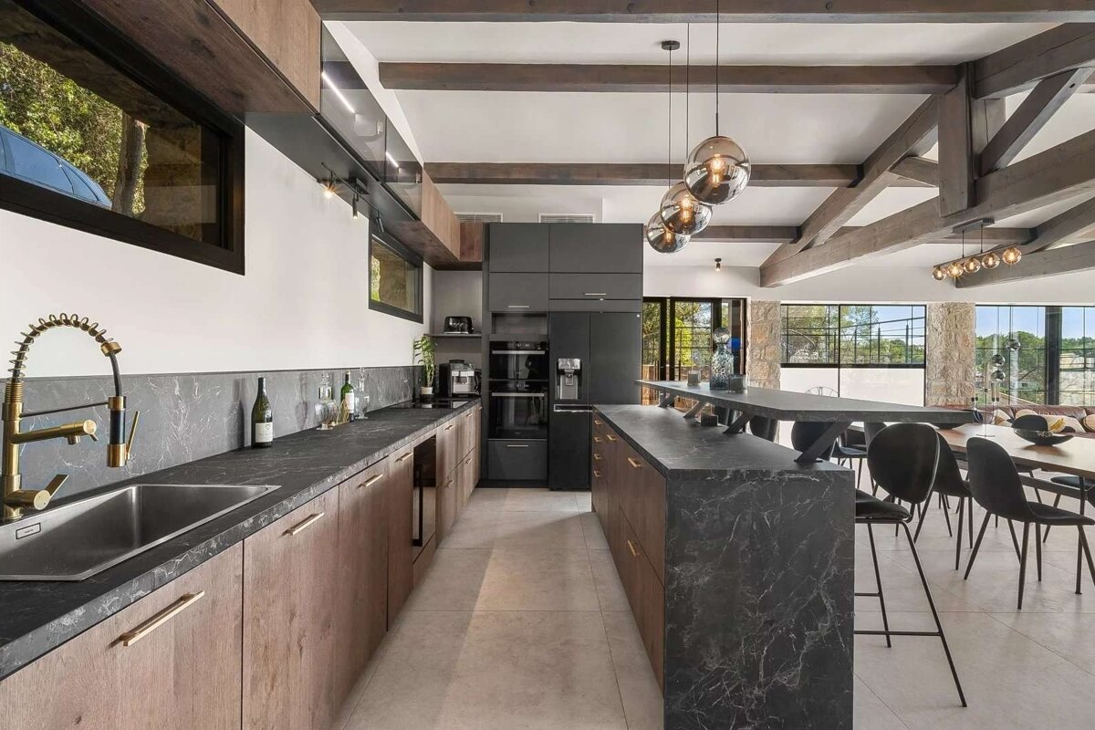 Spacious, modern kitchen with wood cabinets, dark marble countertops, island, and dining area, featuring exposed beams and large windows.