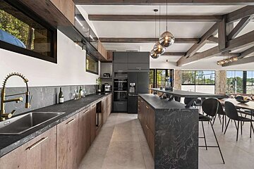 Spacious, modern kitchen with wood cabinets, dark marble countertops, island, and dining area, featuring exposed beams and large windows.
