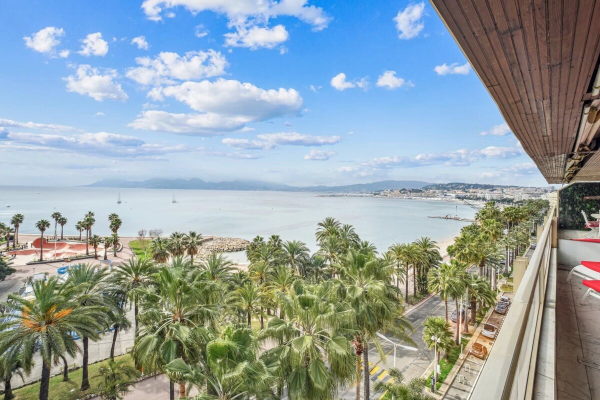 High-angle balcony view of a sunny coastline with a calm blue sea, sandy beach, lush palm trees, and a distant city under a partly cloudy sky.