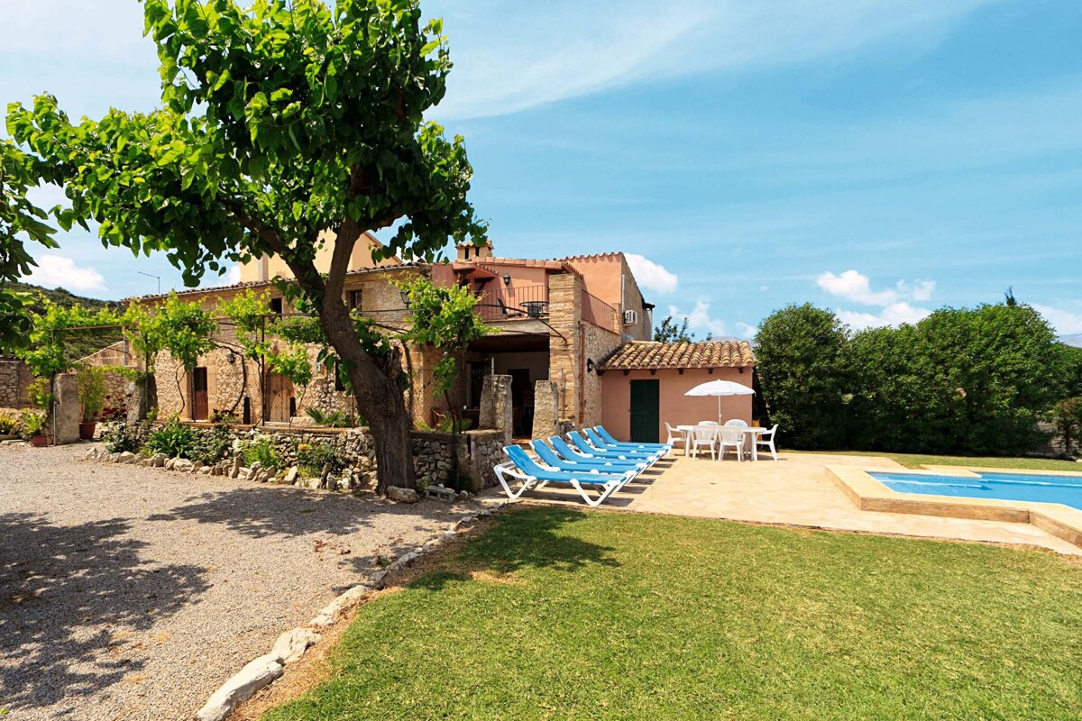 A sunny view of a traditional stone villa with a vibrant green lawn, sparkling pool, blue sun loungers, and an outdoor dining set under a tree.
